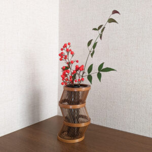 A hand-bent sepia Suruga bamboo single-flower vase with a unique undulating curve, intricate weaving at the base, and an accompanying dark wood stand.