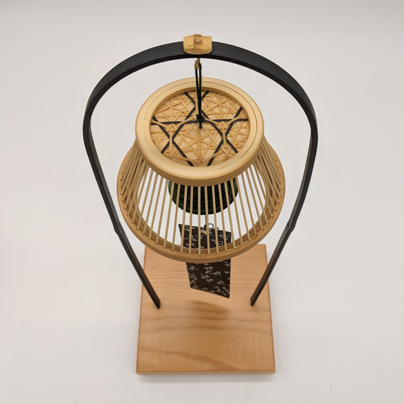 A conical Suruga bamboo lattice cage housing a dark Nambu iron wind bell, suspended from a tall tabletop wooden stand. – view 2